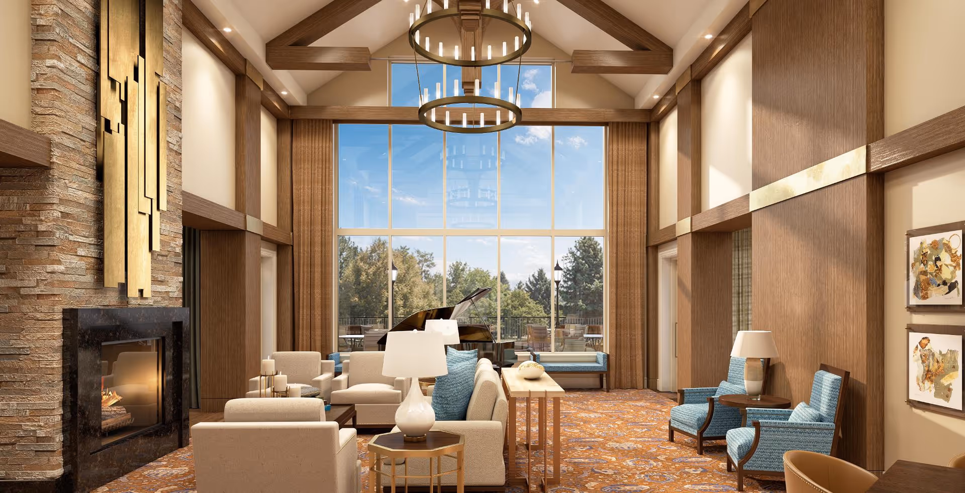 A spacious and elegant living room with high vaulted ceilings and large floor-to-ceiling windows showing a view of trees outside. The room features a stone fireplace on the left, beige sofas and armchairs with blue cushions, a grand piano near the windows, two circular chandeliers hanging from the ceiling, and framed artwork on the right wall. The carpet has a warm, intricate pattern, and there are several table lamps providing soft lighting.