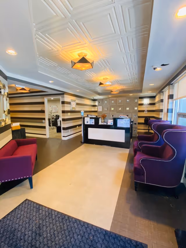 Reception area of South Shore Rehabilitation and Nursing Center featuring a black and white striped wall, a black reception desk with various items on it, two purple armchairs with nailhead trim on the right, a purple sofa on the left, and a patterned ceiling with multiple light fixtures.