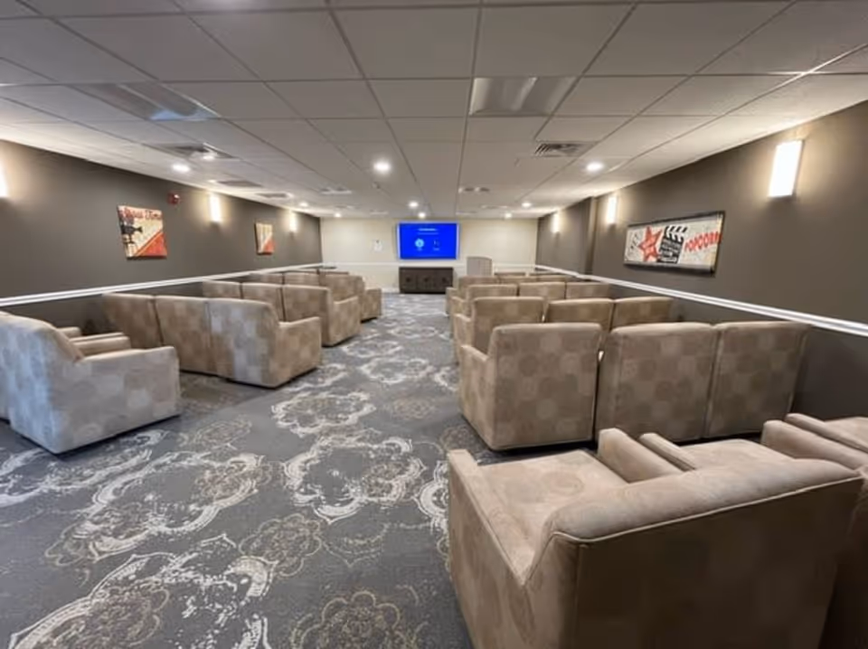 A large room with multiple rows of beige cushioned armchairs arranged facing a wall-mounted flat screen TV. The room has patterned carpet flooring, brown walls with white trim, and several wall-mounted lights. There are framed pictures on the walls, including one with a popcorn theme.