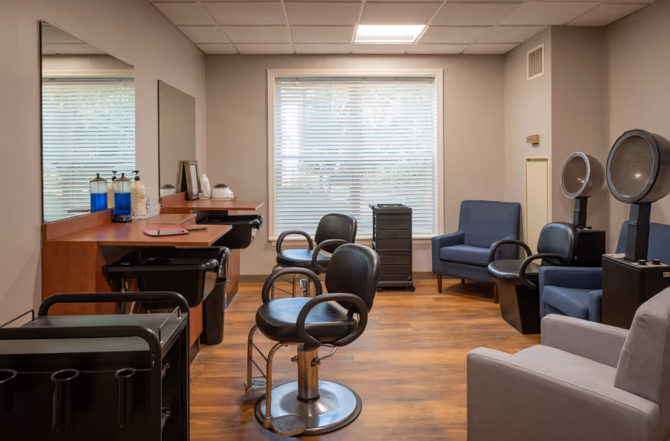 Interior of a salon room with two black salon chairs in front of a wooden counter with mirrors, hair care products, and sinks. There are two blue armchairs and two hair dryer chairs with hooded dryers near a window with blinds. The floor is wooden and the walls are painted beige.
