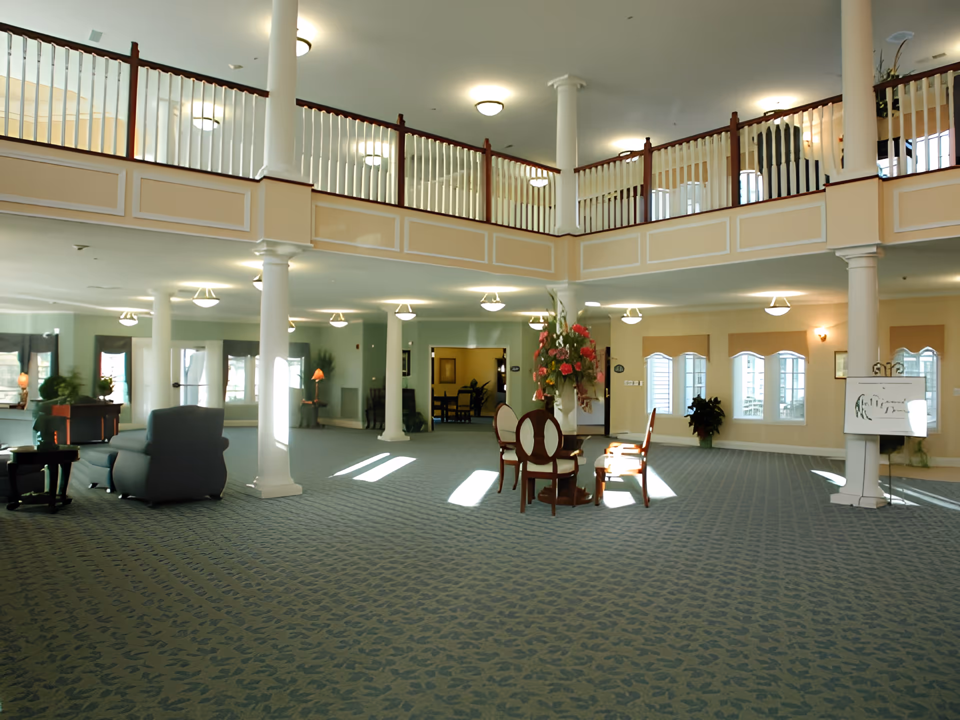 Spacious senior living center lobby with blue patterned carpet, white columns, and a second-floor balcony with wooden railings. The room is furnished with a few chairs and tables, a large floral arrangement in the center, and several windows letting in natural light.