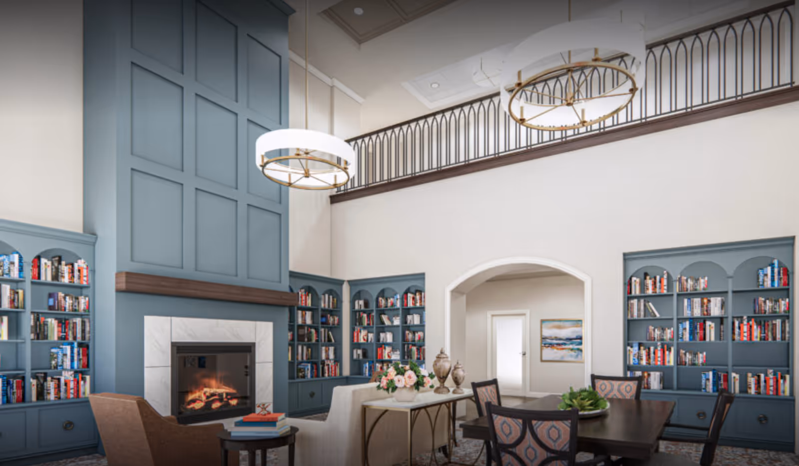 A spacious and elegant living room with high ceilings, featuring a large blue paneled fireplace surrounded by built-in bookshelves filled with books. The room has a cozy seating area with a beige sofa, a brown armchair, a dark wooden table with chairs, and decorative elements including flowers and vases. Two modern circular chandeliers hang from the ceiling, and there is a balcony railing above the fireplace area.