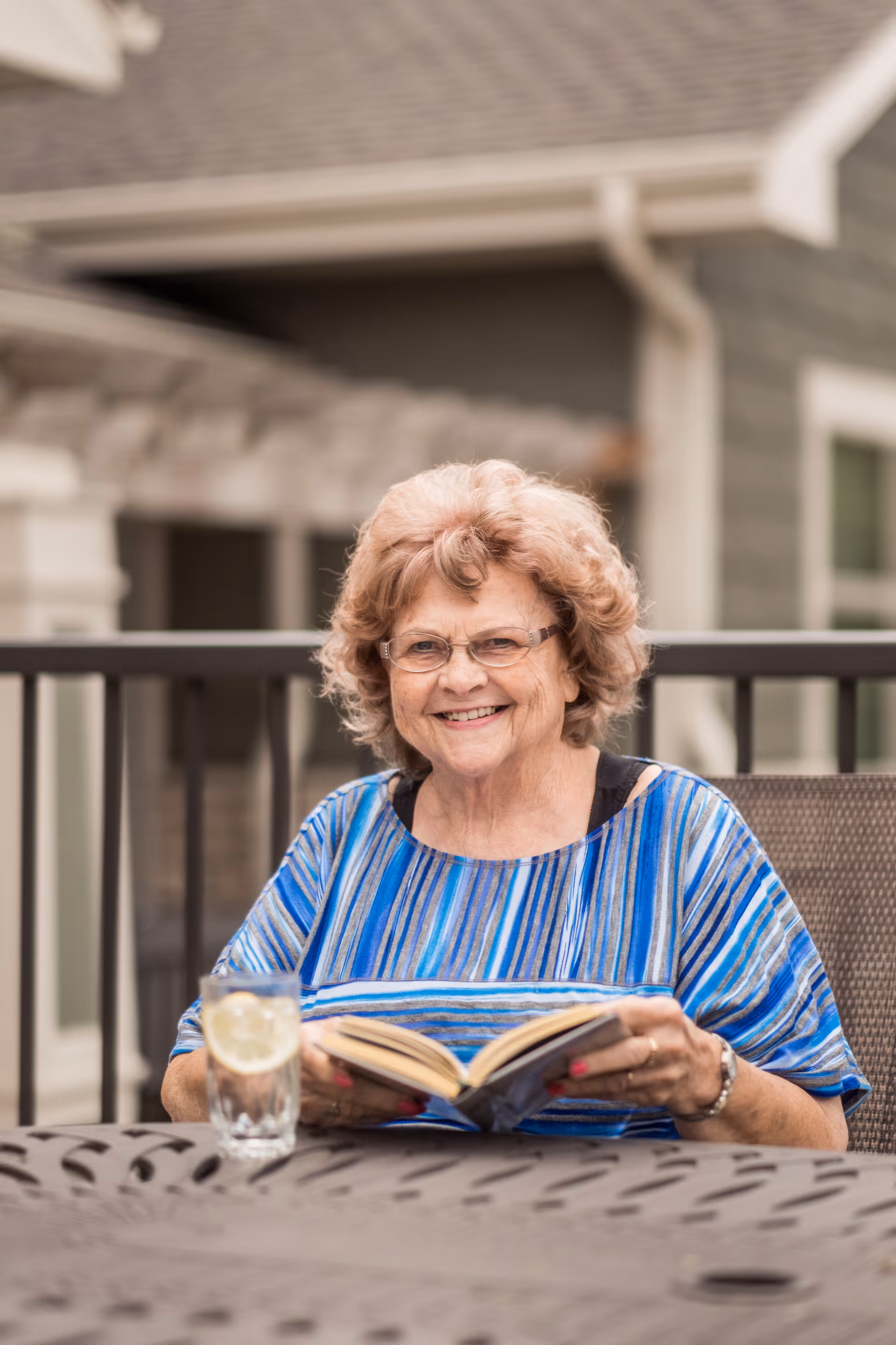 An elderly woman with curly hair and glasses is sitting outdoors at a metal table, smiling while holding an open book. There is a glass of water with lemon on the table in front of her, and a building with a railing is visible in the background.