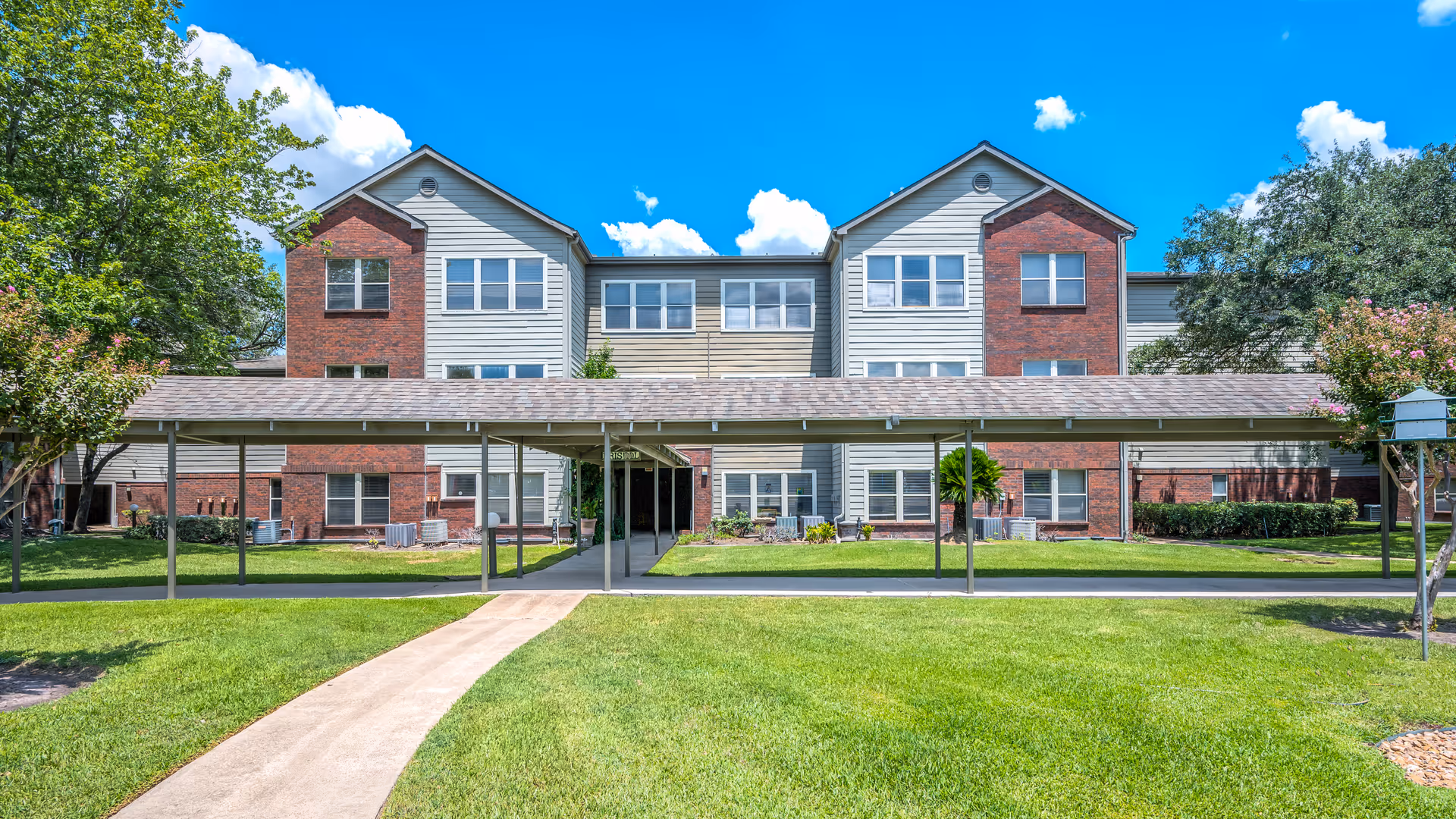 Exterior view of a three-story residential building with a combination of red brick and light-colored siding. A covered walkway leads to the entrance, surrounded by well-maintained green lawns and trees under a bright blue sky with some clouds.