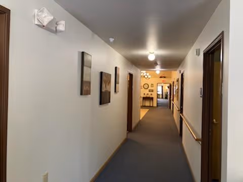 A long indoor hallway with beige walls and carpeted floor. Several framed pictures hang on the left wall, and there are handrails on the right wall. Multiple doorways line both sides of the hallway, and ceiling lights illuminate the space. At the end of the hallway, a room with more furniture and wall decorations is visible.