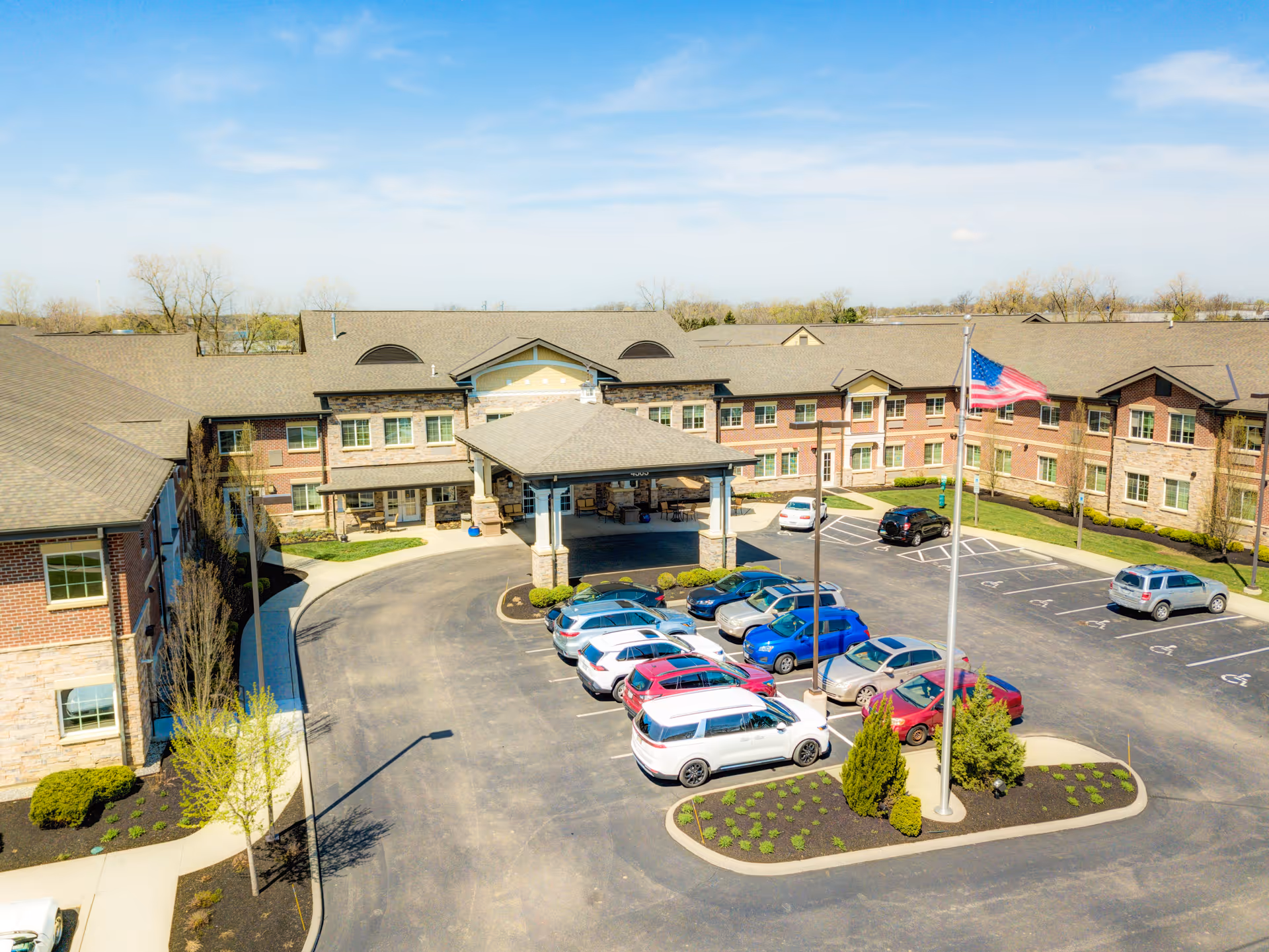 Exterior view of Traditions of Hilliard senior living facility showing a large U-shaped building with brick and stone facade, a covered entrance, a parking lot with several cars, an American flag on a flagpole, and landscaped areas with small trees and bushes under a clear blue sky.