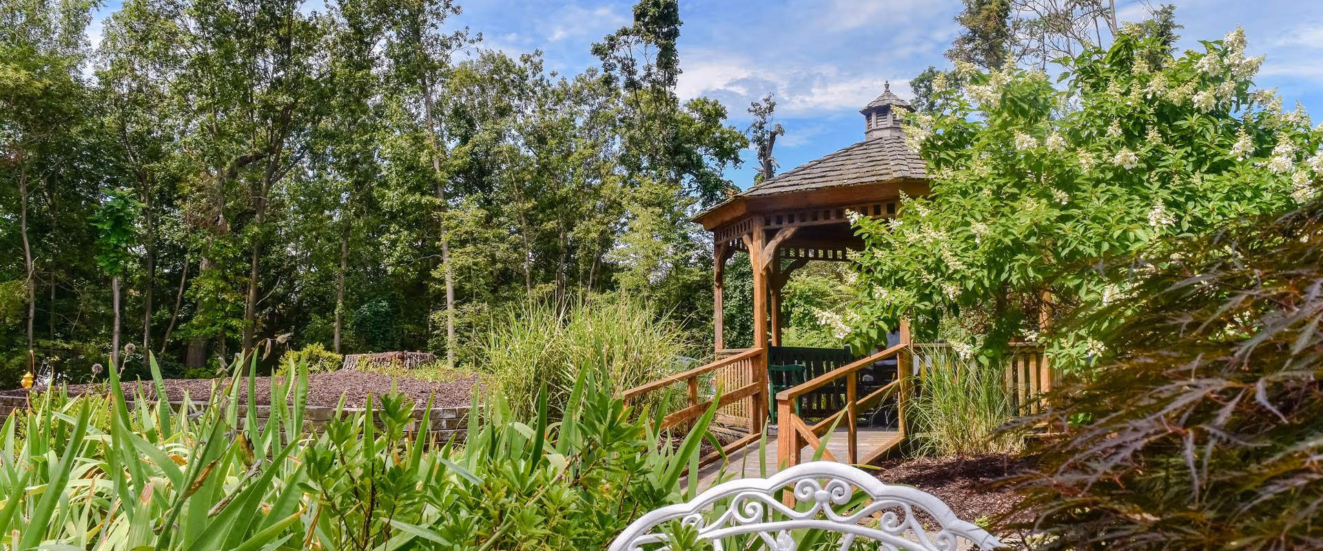A peaceful outdoor garden area with lush green plants, trees, and a wooden gazebo with a shingled roof. In the foreground, there is a white ornate metal bench partially visible. The sky is blue with some clouds.