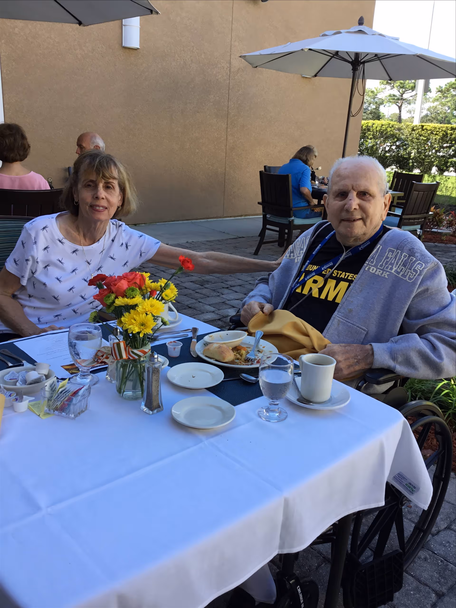 Two elderly people sitting at a table outdoors with a white tablecloth, enjoying a meal. The woman on the left is wearing a white shirt with a dragonfly pattern and holding a red flower, while the man on the right is wearing a gray jacket and a black shirt with the word 'ARMY' visible. The table has plates, cups, glasses of water, and a vase with yellow and red flowers. Other people are seated at tables in the background under umbrellas.