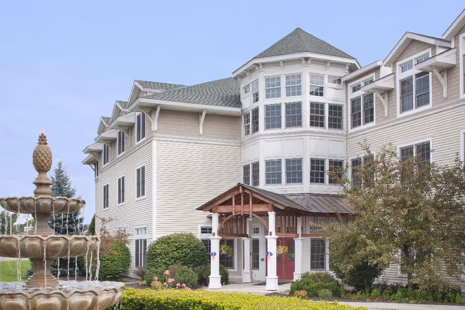 Exterior view of Partridge House, a multi-story senior living facility with beige siding and green roofing. The entrance features a covered porch with white pillars and wooden beams. In the foreground, there is a decorative stone fountain with water flowing, surrounded by well-maintained shrubs and greenery.