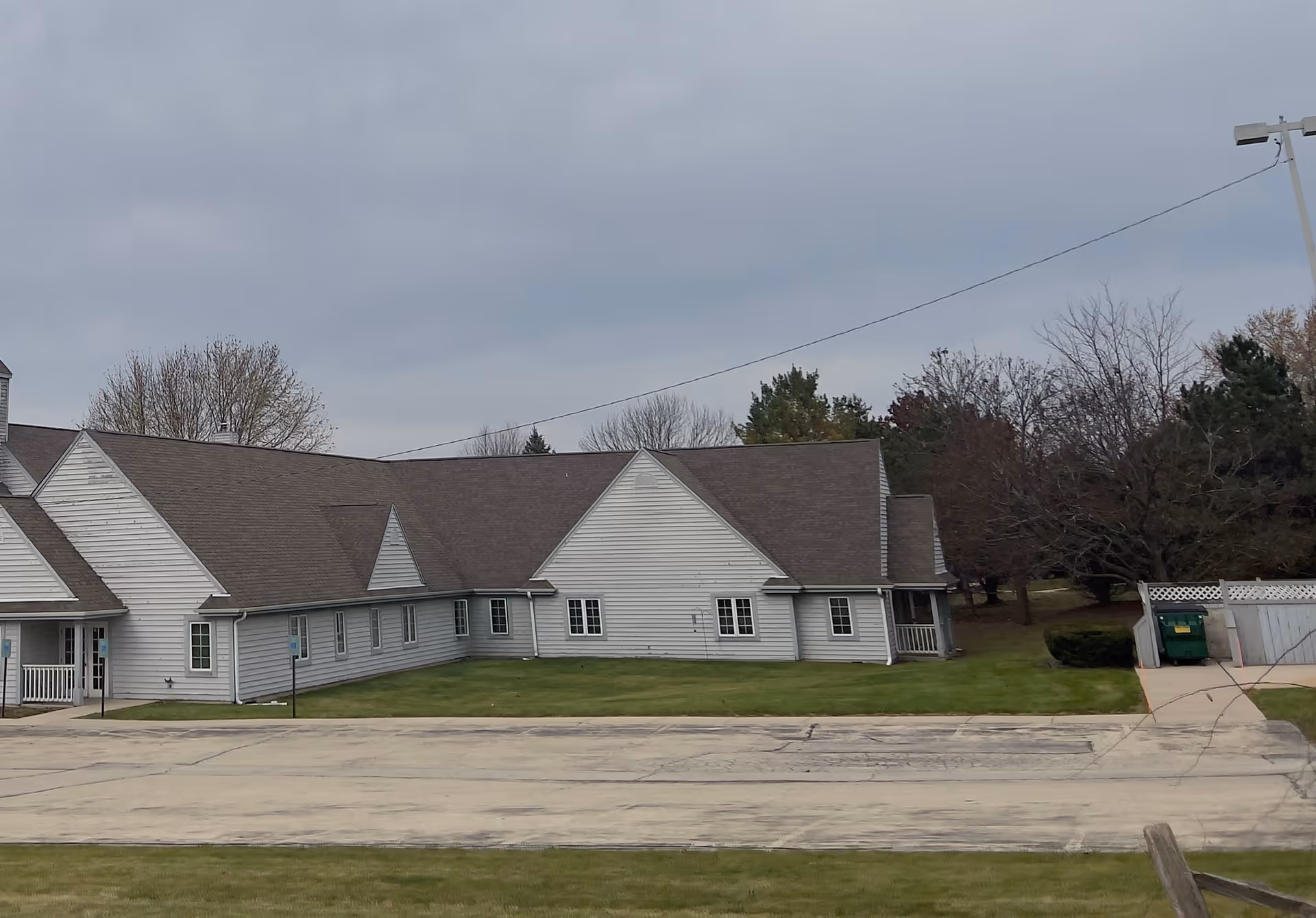 Exterior view of a single-story building with white siding and a dark shingled roof, surrounded by grass and trees under a cloudy sky. There is a paved area in front and a green dumpster near the right side of the building.