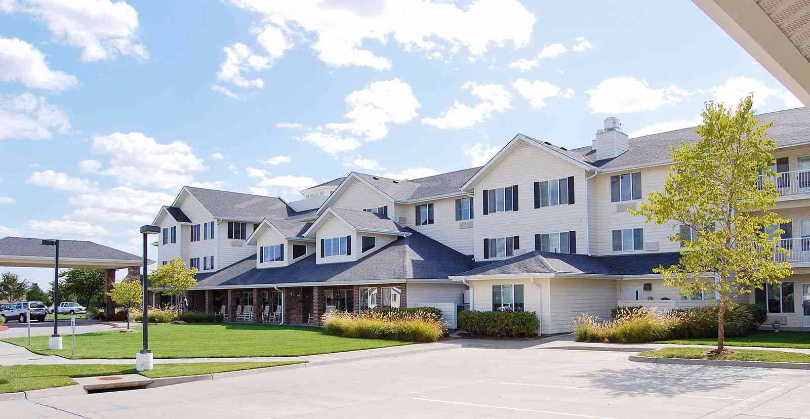 Exterior view of a multi-story senior living facility building with white siding and black shutters. The building has a covered entrance with rocking chairs on the porch, surrounded by green grass, small trees, and a clear blue sky with scattered clouds.