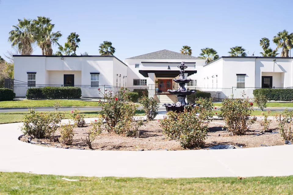 Front exterior view of a single-story white building with a central entrance marked with the letter A, surrounded by a garden area with bushes and a black multi-tiered fountain in the foreground, palm trees in the background, and a clear blue sky.