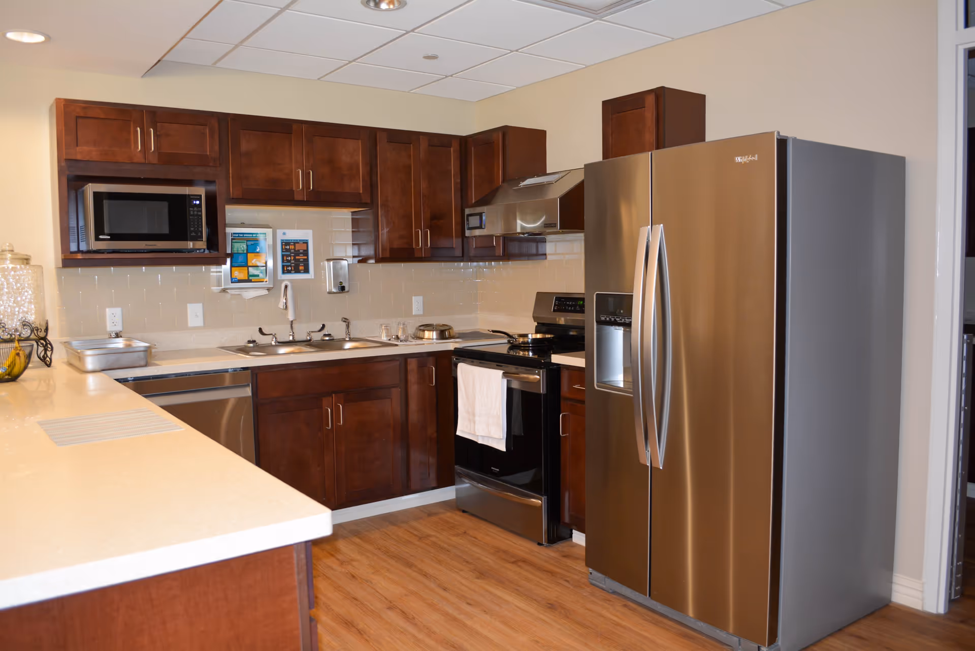 A modern kitchen with wooden cabinets, a stainless steel refrigerator, stove with a white towel hanging on the handle, microwave, double sink, and light-colored countertops. The floor is wooden, and the walls are painted beige with a tiled backsplash.