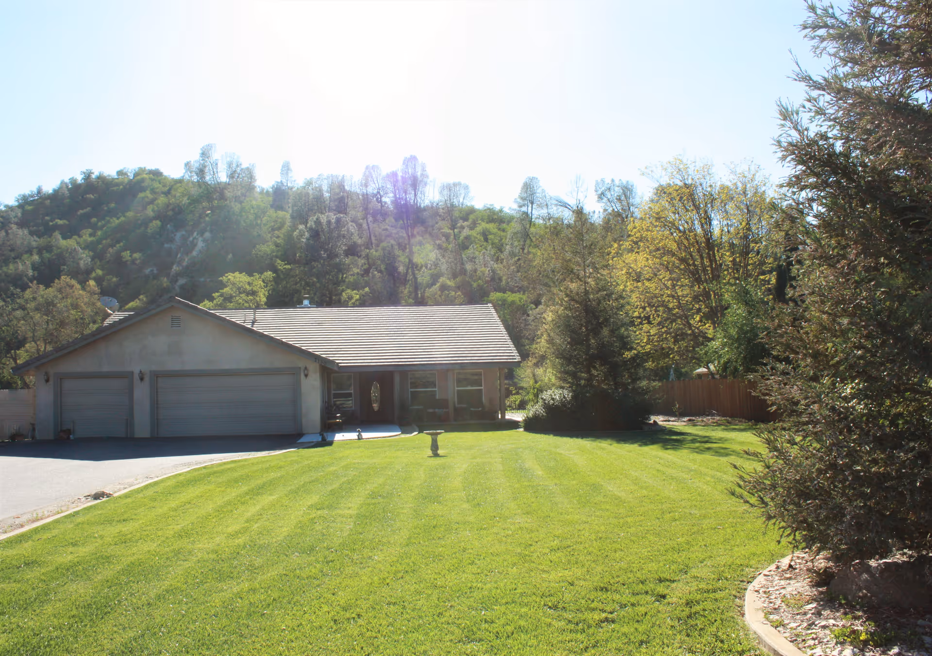 A single-story house with a three-car garage, surrounded by a well-maintained green lawn and trees, with a wooded hillside in the background under a bright sky.