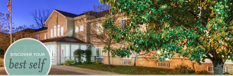 Exterior view of a two-story senior living facility building at dusk with lights on inside. There is a large tree with green leaves in the foreground and an American flag on a flagpole to the left. A circular graphic in the bottom-left corner reads 'DISCOVER YOUR best self'.