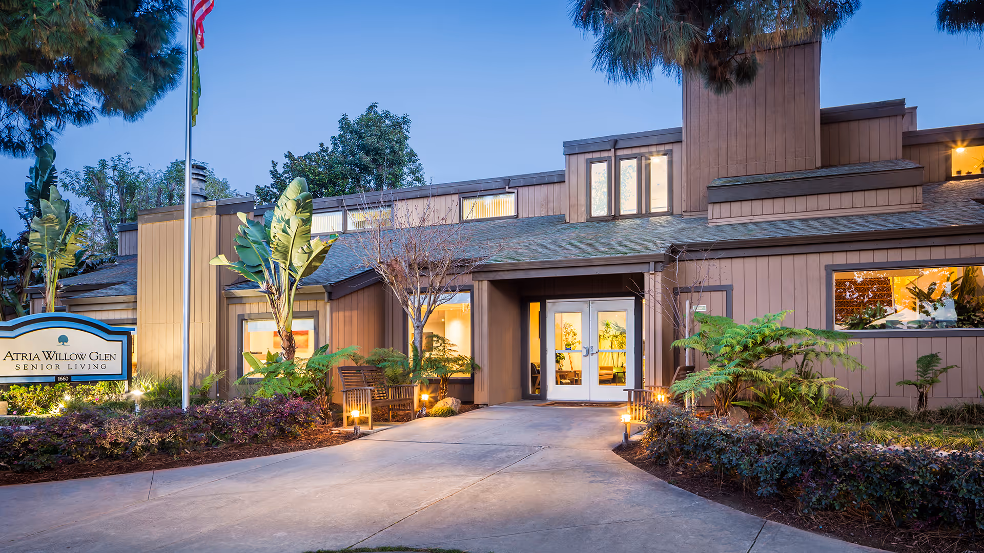 Exterior view of Atria Willow Glen senior living facility at dusk, showing the entrance with double glass doors, surrounding landscaping with plants and trees, a bench, and a sign with the facility name.