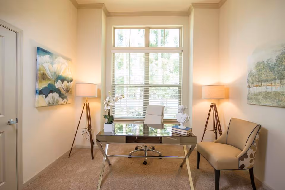 A bright office space with a large window letting in natural light. The room features a modern glass desk with a white office chair behind it. On the desk are a white orchid plant, a small bust sculpture, and a stack of books. There are two tripod floor lamps with beige lampshades on either side of the desk. A beige upholstered chair with a floral pattern on the side is positioned to the right. The walls are decorated with two paintings, one with white flowers on the left and a landscape on the right.