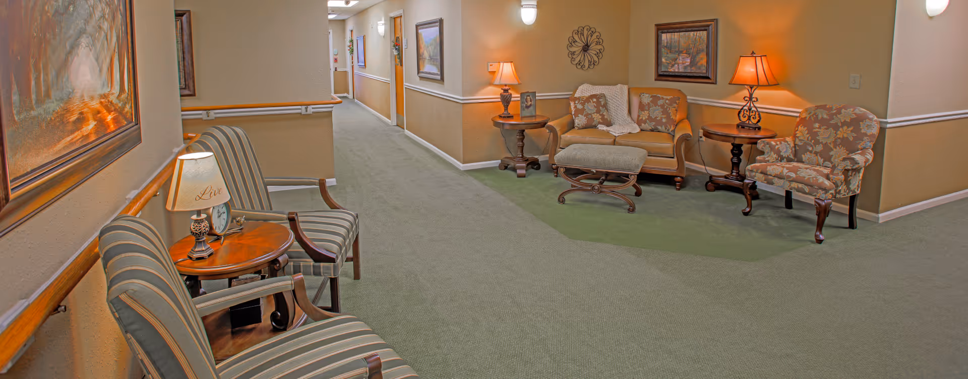 A well-lit hallway in a senior living facility with green carpet and beige walls. The hallway features a seating area with a floral-patterned armchair, a small sofa with cushions, an ottoman, and side tables with lamps. There are framed paintings and decorative wall art on the walls, and handrails along the hallway.