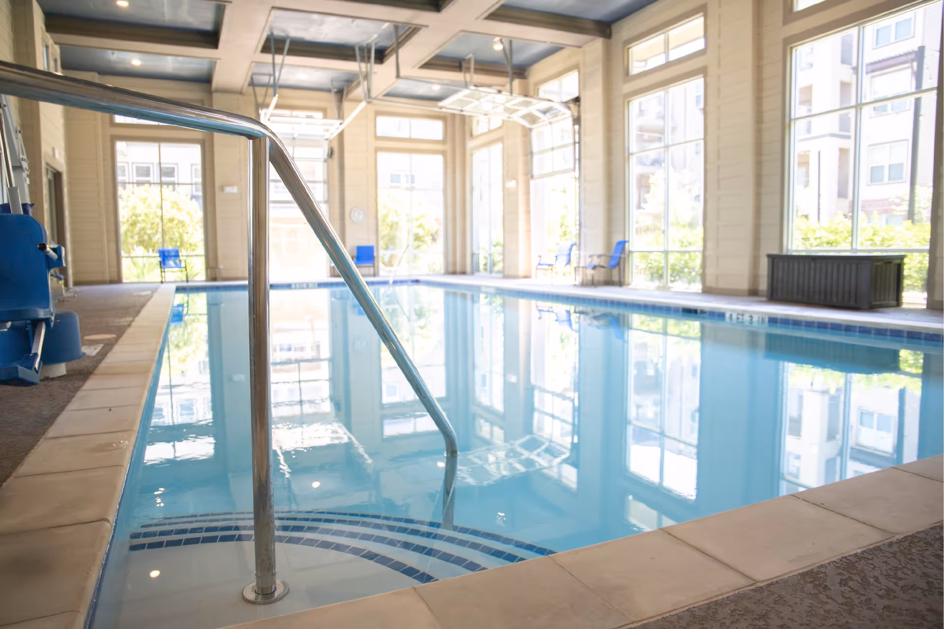 Indoor swimming pool with clear blue water, metal handrail, large windows letting in natural light, and blue chairs along the far side of the pool area.