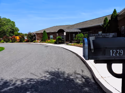 Curved driveway leading to the entrance of Bedford County Nursing Home, a single-story brick building with landscaping and a mailbox numbered 1229 in the foreground.