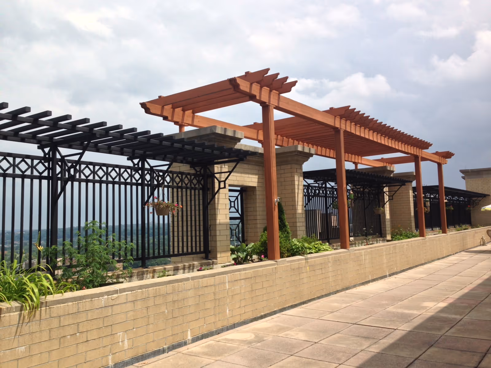 Outdoor terrace area with a tiled floor, beige brick planter walls, black metal fencing, and wooden pergolas overhead. Some plants and hanging flower pots are visible along the planter walls under a cloudy sky.