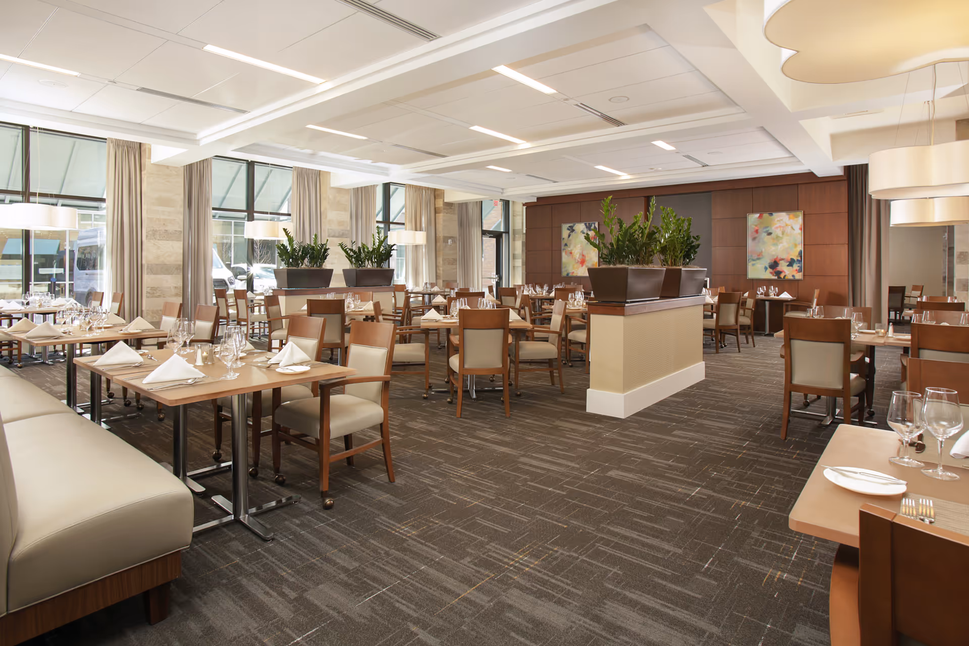 A spacious and well-lit dining room with multiple tables set with white napkins, glassware, and silverware. The room features beige cushioned chairs, large windows with curtains, potted plants on dividers, modern ceiling lights, and abstract paintings on the wooden-paneled walls.