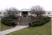 Exterior view of a single-story building with a central entrance accessed by a wide staircase flanked by bushes and trees without leaves, with a pathway leading up to the stairs.