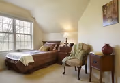 A cozy bedroom with a single bed covered in a brown quilt and a beige throw blanket. Next to the bed is a wooden nightstand with a lamp and a small clock. There is an upholstered armchair with a green throw draped over it, and a wooden side table with a red vase. A framed picture hangs on the wall above the side table. A large window lets in natural light.