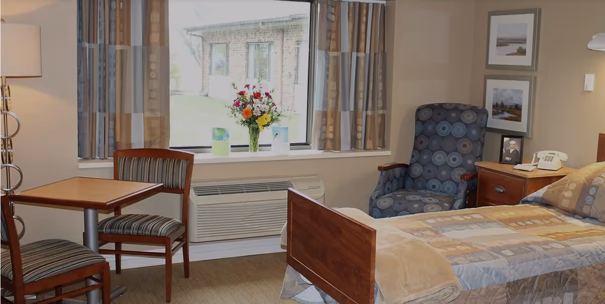 A cozy bedroom in a senior living facility featuring a single bed with a patterned quilt, a wooden nightstand with a telephone and framed photo, a comfortable upholstered armchair with a circular pattern, a small wooden table with two striped chairs, a window with patterned curtains, and a vase of colorful flowers on the windowsill.