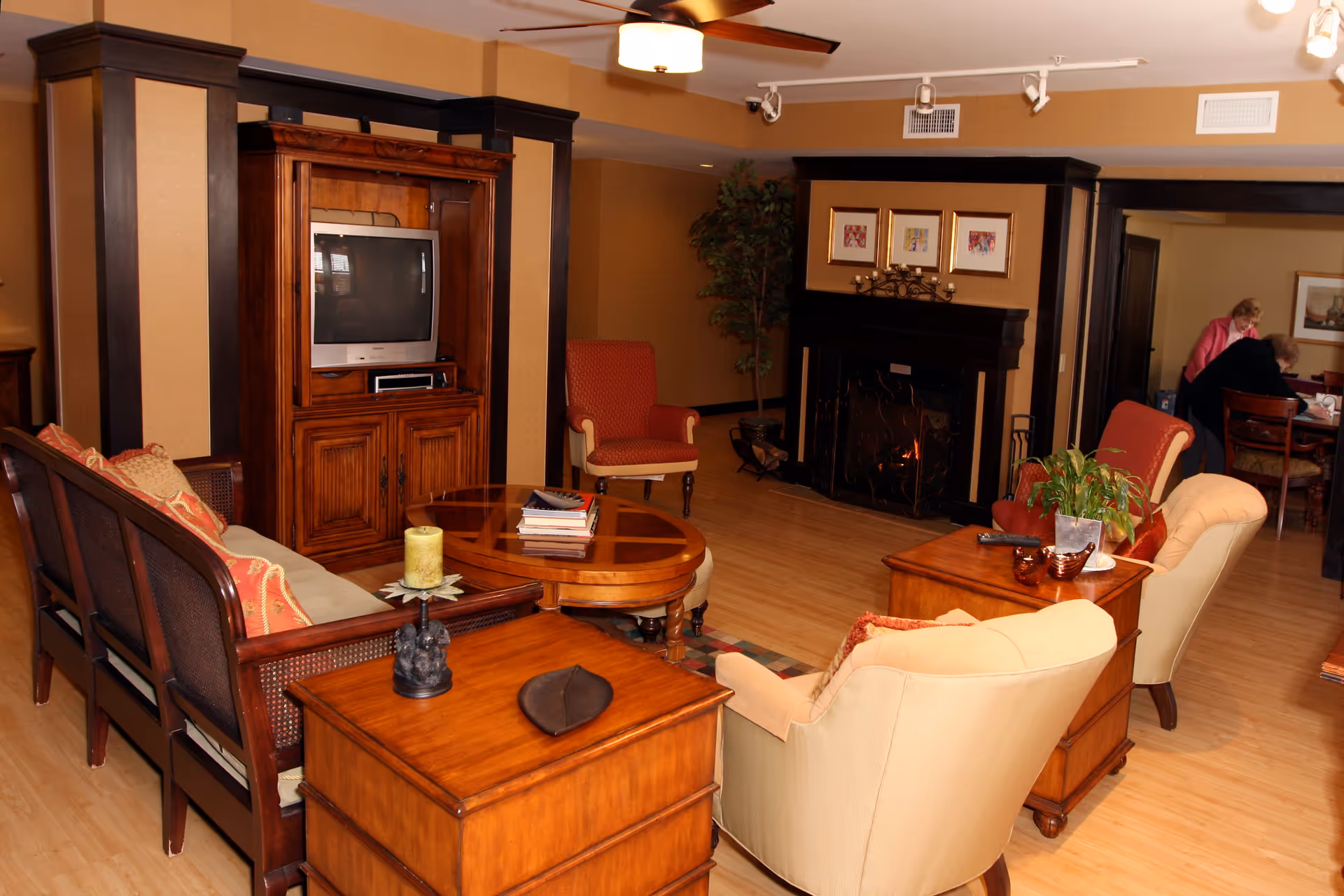 A cozy living room with wooden furniture including a TV cabinet with a television, a coffee table with books, armchairs, and a sofa with cushions. There is a fireplace with framed artwork above it and a ceiling fan with a light. In the background, two people are seated at a table in an adjoining room.