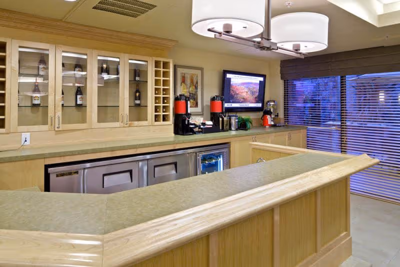 Interior view of a kitchen or serving area with a light wood counter and cabinetry. The counter has a smooth surface with a raised wooden edge. Behind the counter, there are glass-front cabinets displaying bottles, a wine rack, two red beverage dispensers, a coffee maker, and a small refrigerator. A flat-screen TV is mounted on the wall above the counter, and large windows with horizontal blinds are visible on the right side.