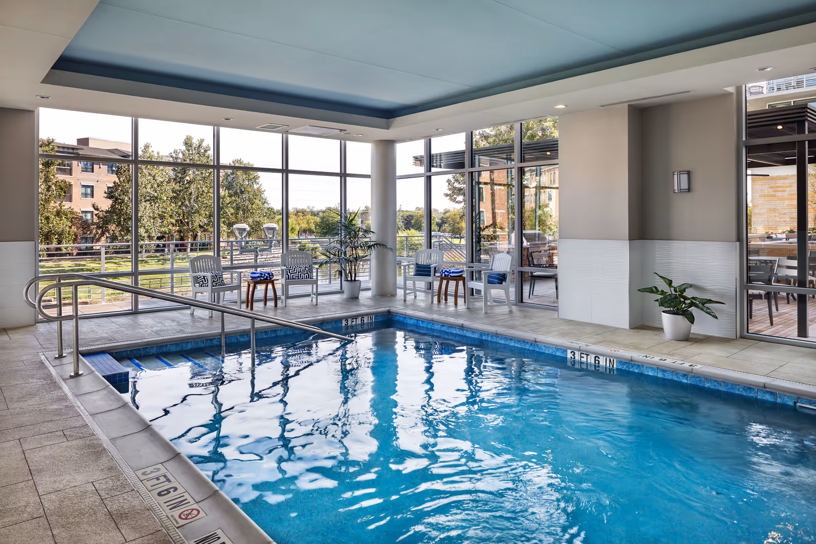 Indoor swimming pool area with clear blue water, surrounded by large floor-to-ceiling windows showing an outdoor view with trees and buildings. There are white chairs with blue cushions and small wooden tables along the poolside, as well as potted plants. The ceiling has a recessed blue section with lighting.