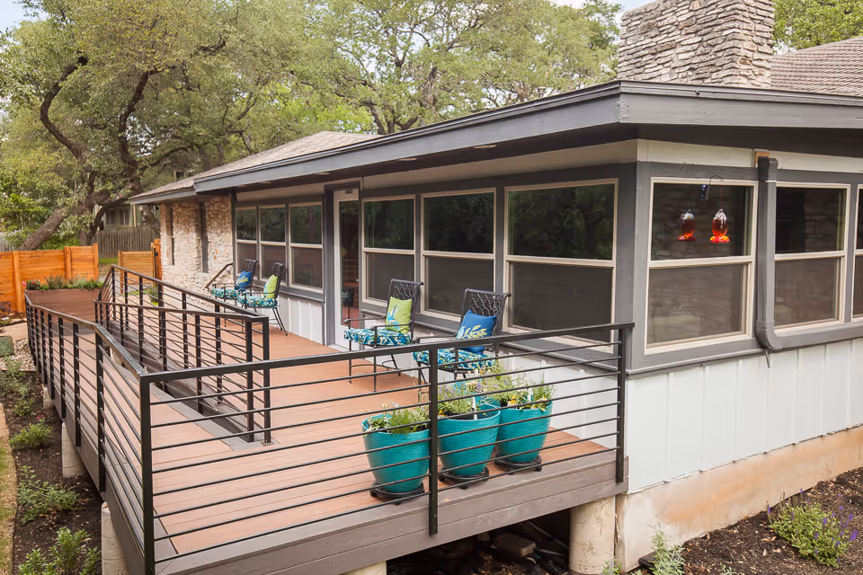 Outdoor wooden deck attached to a building with black metal railings, several turquoise planters with green plants, and black metal chairs with blue and green cushions. The building has large windows and a stone chimney, surrounded by trees and greenery.