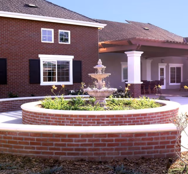 Outdoor courtyard area of a senior living facility featuring a circular brick planter with greenery and a tiered water fountain in the center, surrounded by a paved walkway. The background shows a brick building with windows and a covered patio area with columns and outdoor seating.