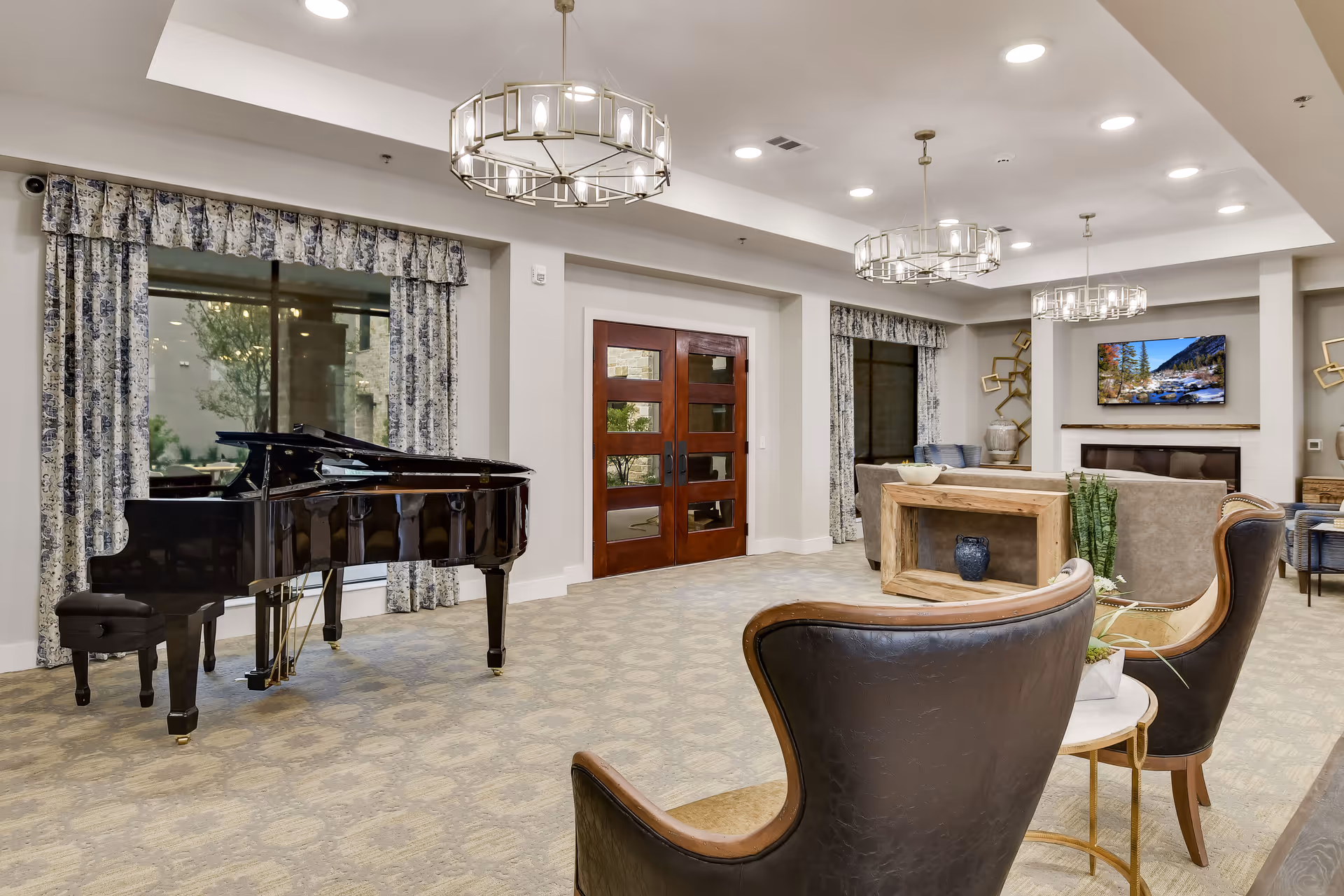 A spacious and elegant living room area in SoCo Village featuring a black grand piano near large windows with patterned curtains. The room has beige carpeting, modern chandeliers, comfortable seating including leather chairs and sofas, a wooden console table with decorative items, and a wall-mounted TV above a fireplace.