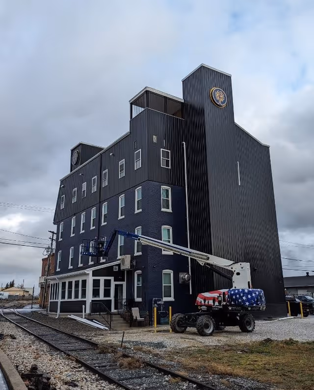 A multi-story dark-colored building with a modern design, featuring a logo with a tree on the upper part of the structure. In front of the building, there is a construction lift with an American flag design on its base. The building is situated next to railroad tracks under a cloudy sky.