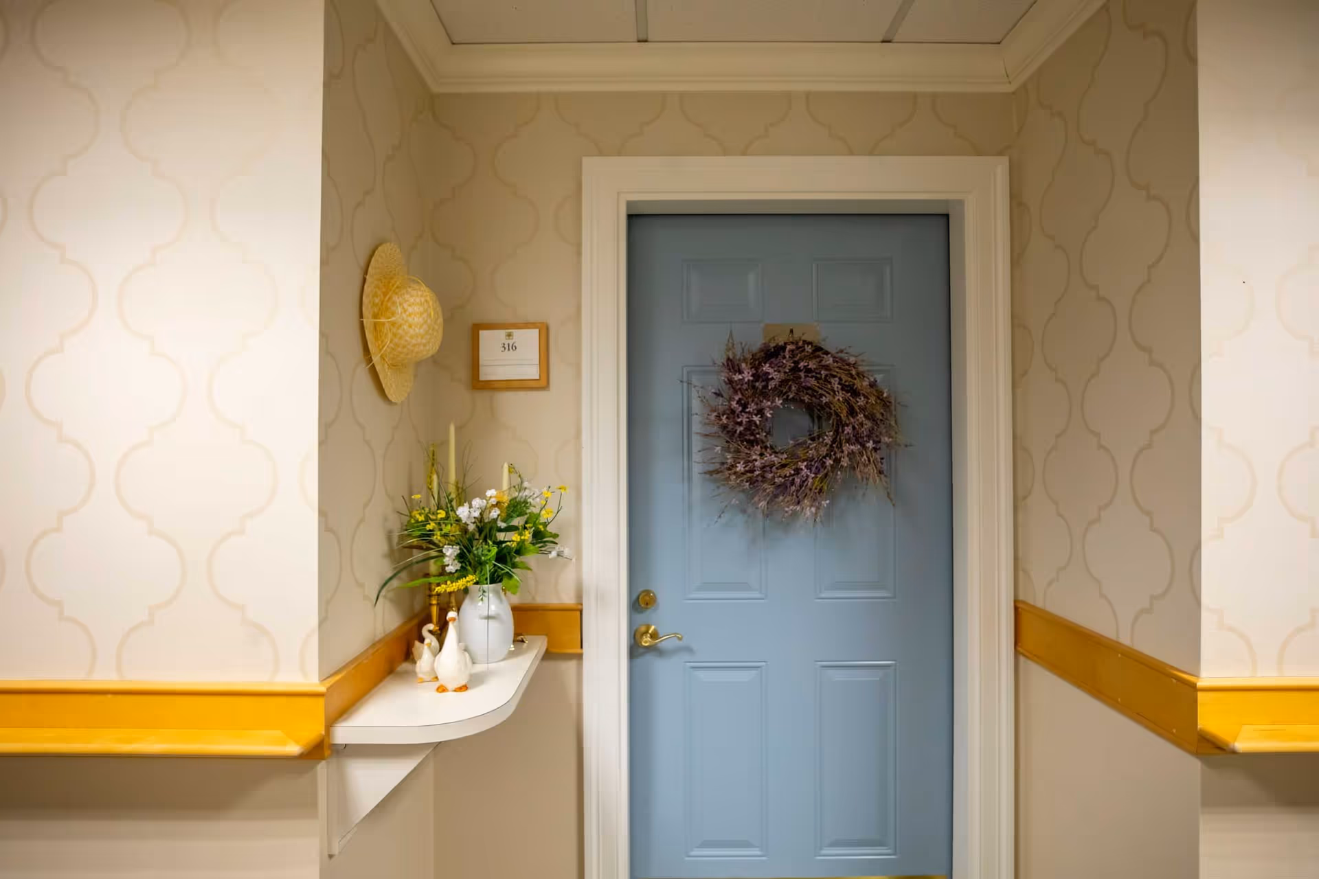 A hallway corner in Edelweiss Village featuring a closed light blue door decorated with a purple floral wreath. To the left of the door, a small white shelf holds a vase with yellow and white flowers and three small ceramic ducks. A straw hat hangs on the wall above the shelf, and a room number plaque labeled '316' is mounted on the wall.
