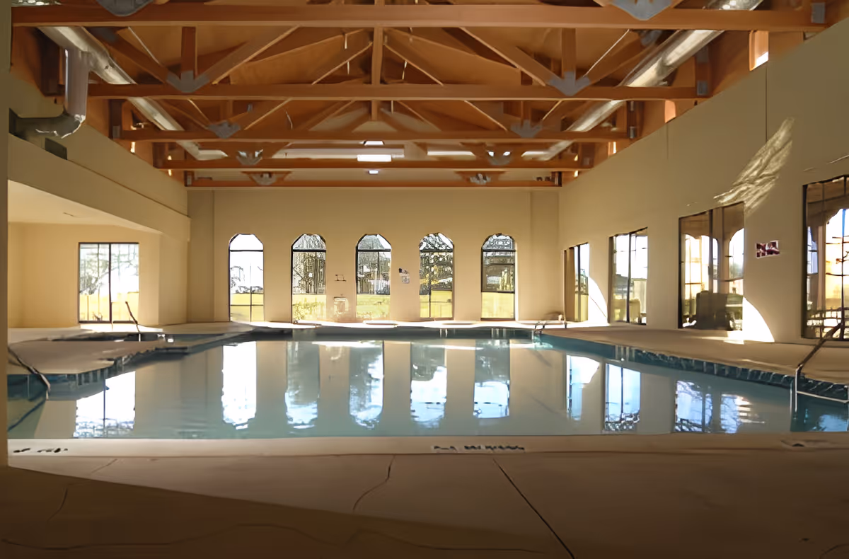 Indoor swimming pool with clear blue water, surrounded by beige walls and large windows letting in natural light. The ceiling features exposed wooden beams and metal supports. The pool area has steps and handrails for entry.