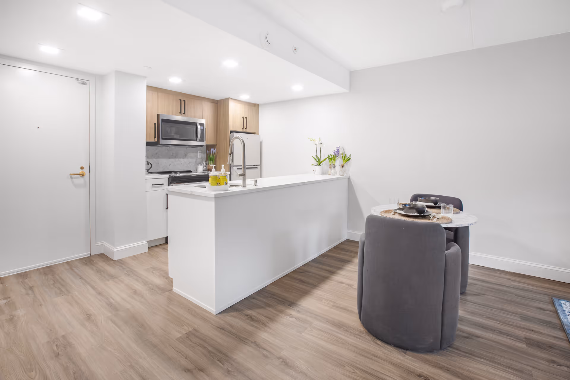 Modern kitchen area with a white island countertop featuring a sink and faucet, wooden cabinets, stainless steel microwave, refrigerator, and stove. Adjacent to the kitchen is a small round dining table set with two gray upholstered chairs and tableware. The room has light wood flooring and white walls.