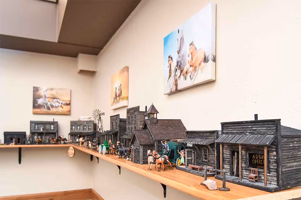 A detailed miniature model of a western town displayed on a wooden shelf mounted on a beige wall. The model includes small buildings, horse-drawn carriages, and figurines of people. Above the shelf, there are paintings of running horses hanging on the wall.