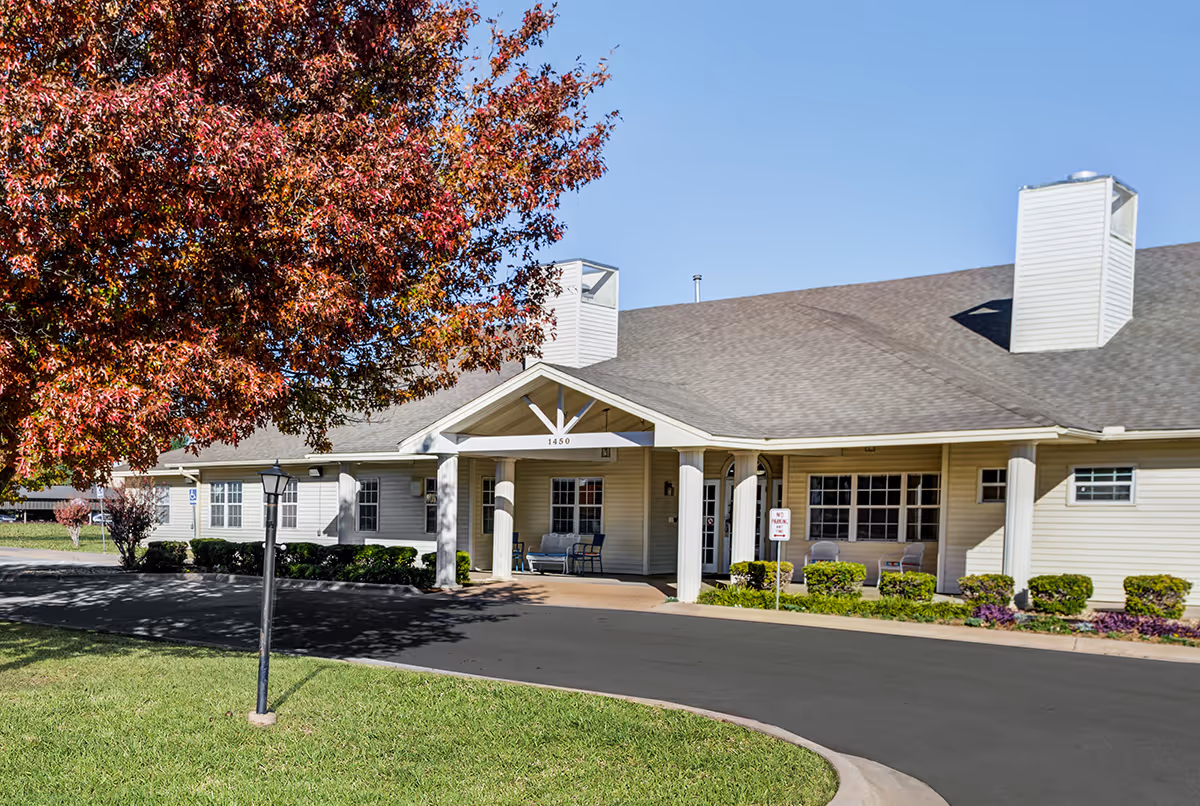 Exterior view of a single-story senior living facility building with beige siding and a brown shingled roof. The entrance has a covered porch supported by white columns, with benches on either side. There is a large tree with red and orange autumn leaves on the left side of the image, a lamp post near the driveway, and well-maintained green grass and shrubs surrounding the building under a clear blue sky.