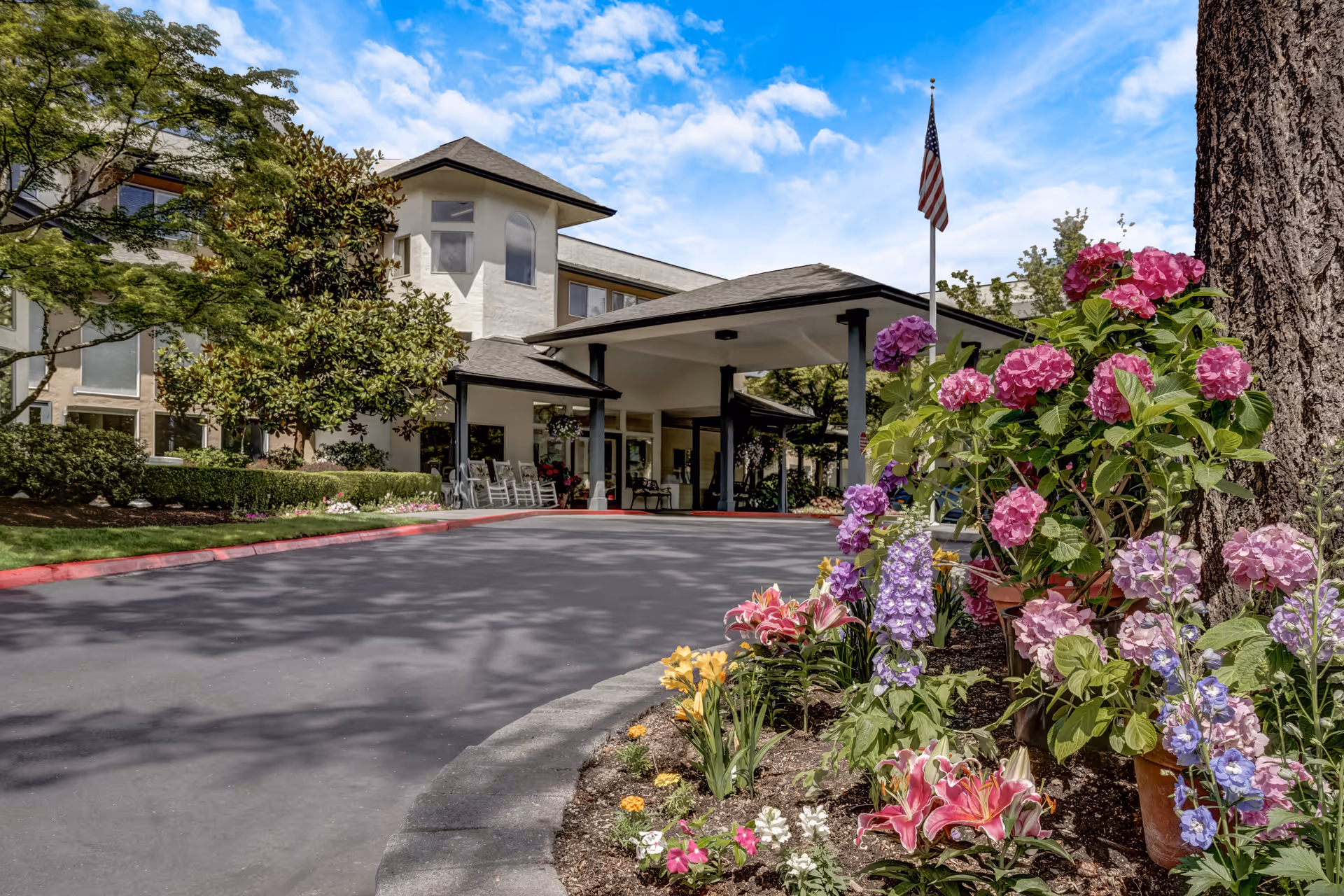 Exterior view of Solista Bellevue senior living facility with a driveway leading to a covered entrance. The building is surrounded by green trees and colorful flowers including pink, purple, and yellow blooms. An American flag is visible near the entrance under a partly cloudy blue sky.