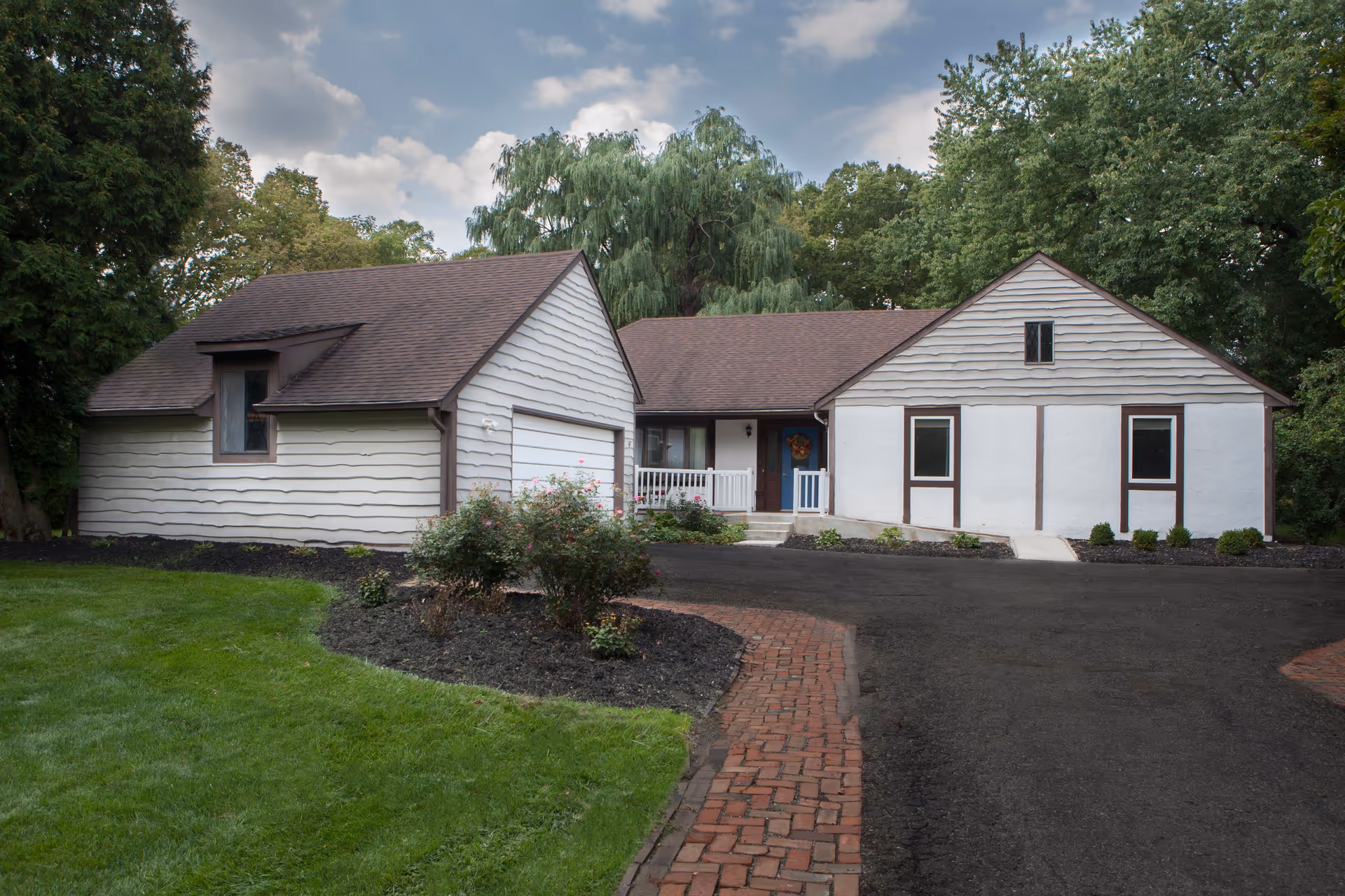 Front exterior view of a single-story house with white siding and brown trim, a brown shingled roof, a small porch with white railing, surrounded by green trees and bushes, with a brick walkway and asphalt driveway in front.