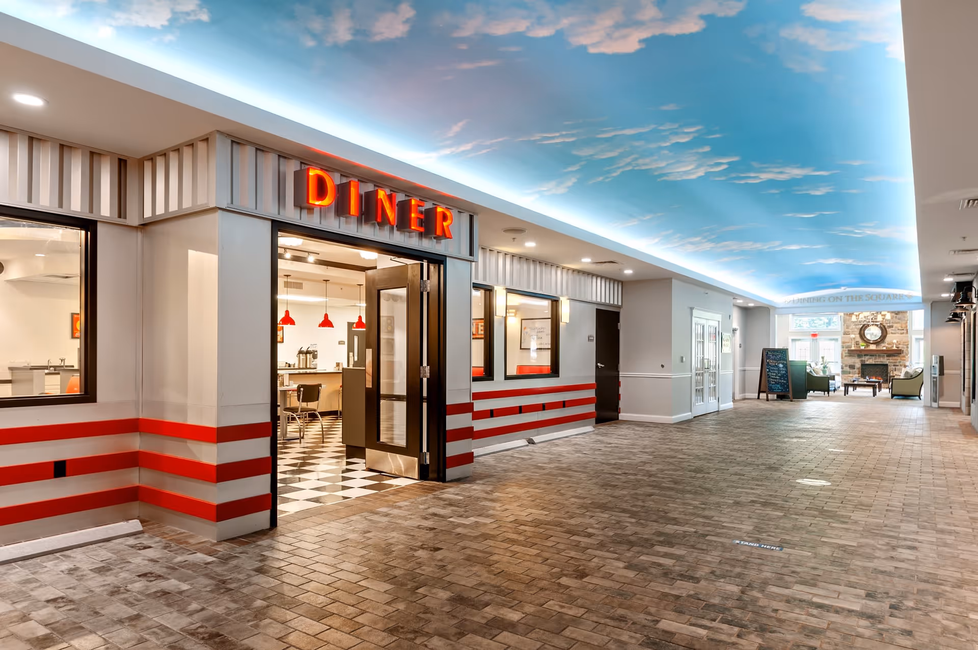 Interior view of a senior living facility hallway with a diner entrance on the left featuring a red neon sign that says DINER. The diner has a retro style with checkered black and white flooring and red pendant lights. The hallway has a ceiling painted with a blue sky and clouds, and at the far end, there is a seating area with green chairs and a stone fireplace.