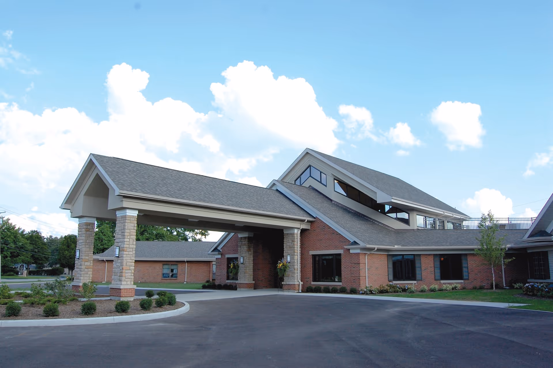 Exterior view of the Franciscan Care Center building featuring a covered entrance with stone pillars, brick walls, multiple windows, and a sloped roof under a partly cloudy blue sky.