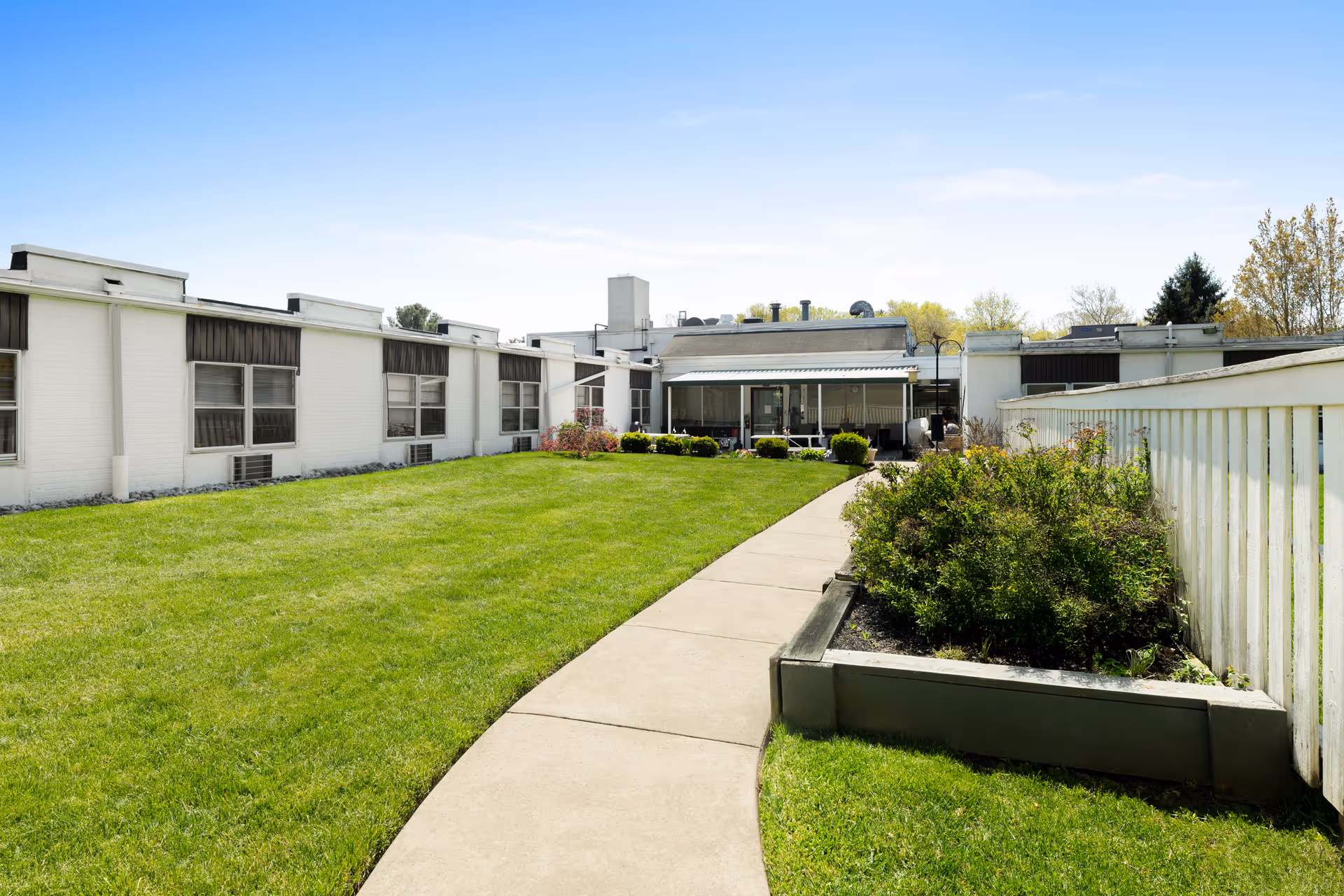 A sunny outdoor courtyard area at Heritage Pointe Rehabilitation and Healthcare Center featuring a curved concrete walkway, green grass lawn, bushes, and a white building with multiple windows and a covered patio area in the background.