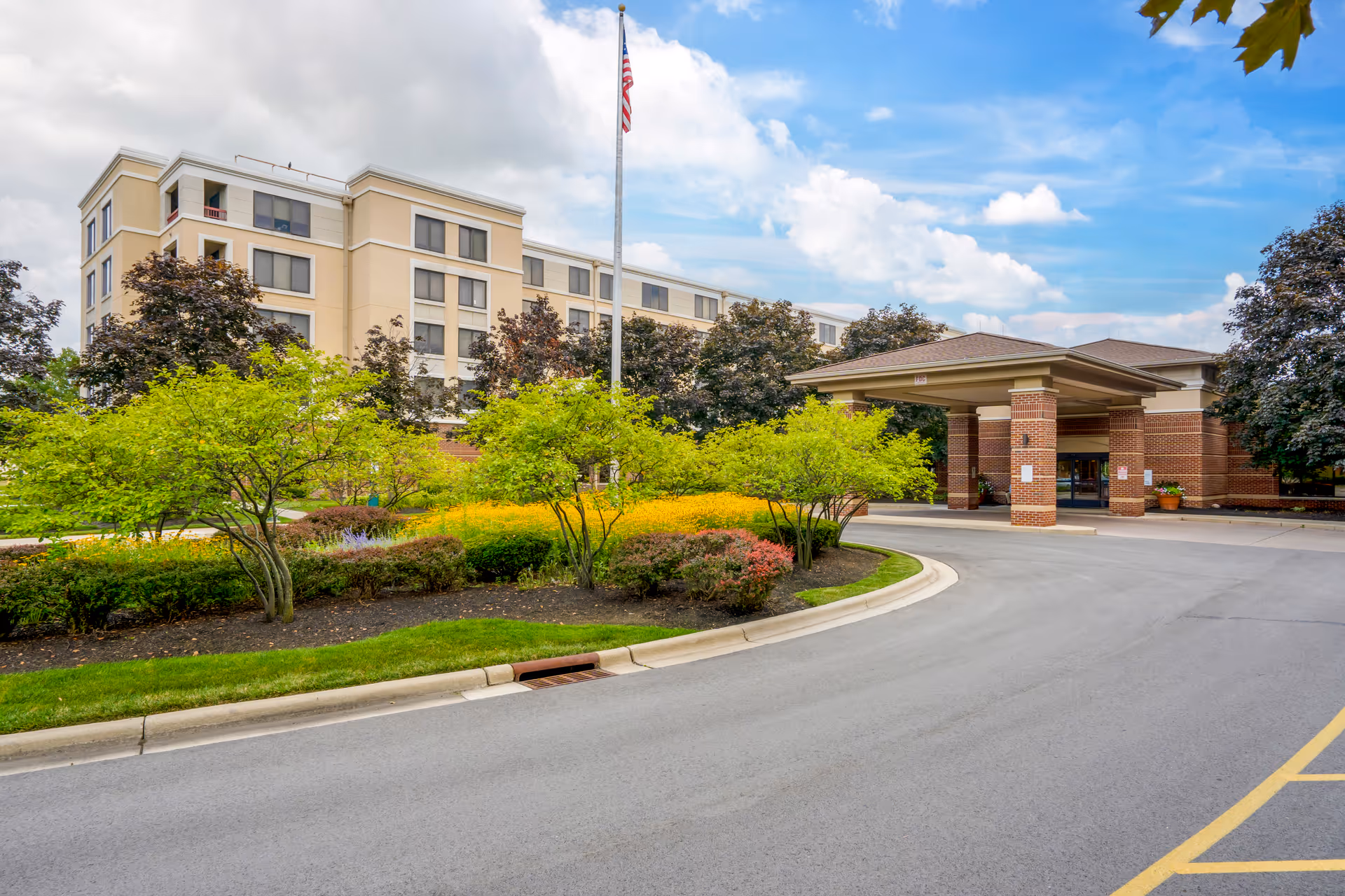 Exterior view of a senior living facility with a multi-story beige building, landscaped greenery, and a covered entrance with brick pillars. An American flag flies on a flagpole in front of the building under a partly cloudy sky.