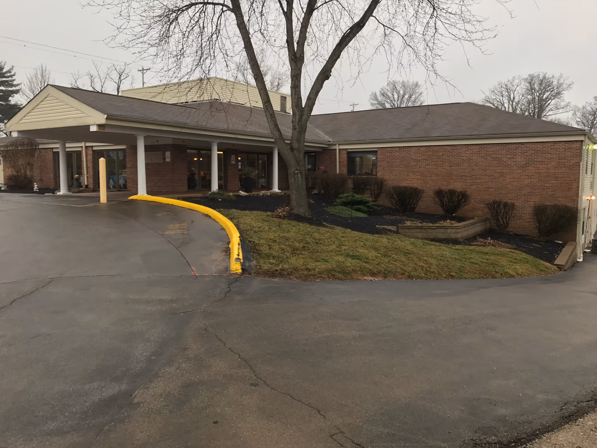 Exterior view of a single-story brick building with a covered entrance supported by white columns. There is a curved driveway with a yellow-painted curb leading to the entrance. Leafless trees and some shrubbery are visible around the building under an overcast sky.