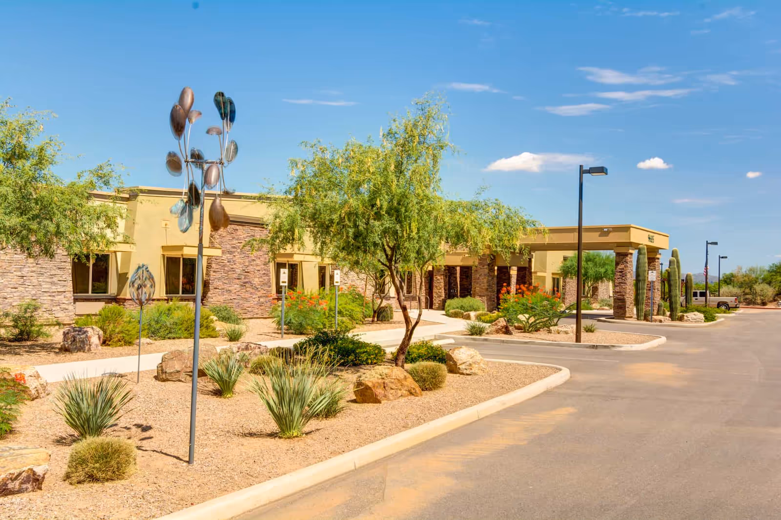 Exterior view of Catalina Springs Memory Care facility showing a single-story building with stone and beige walls, desert landscaping with rocks, shrubs, and small trees, a paved driveway, and a clear blue sky.