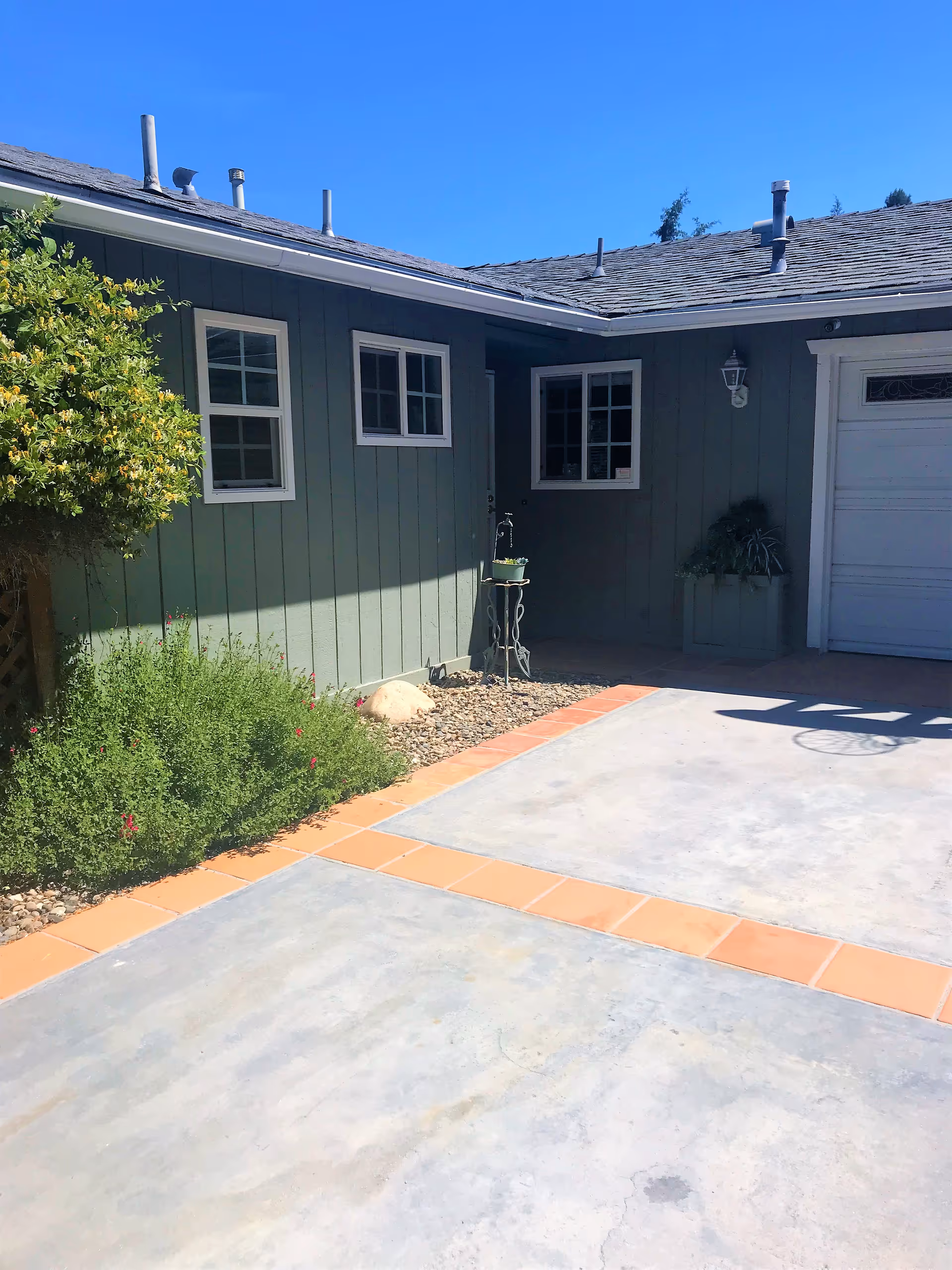 Exterior view of a single-story building with green siding, three white-framed windows, a garage door, and a concrete driveway bordered with orange tiles. There are plants and bushes along the side of the building under a clear blue sky.